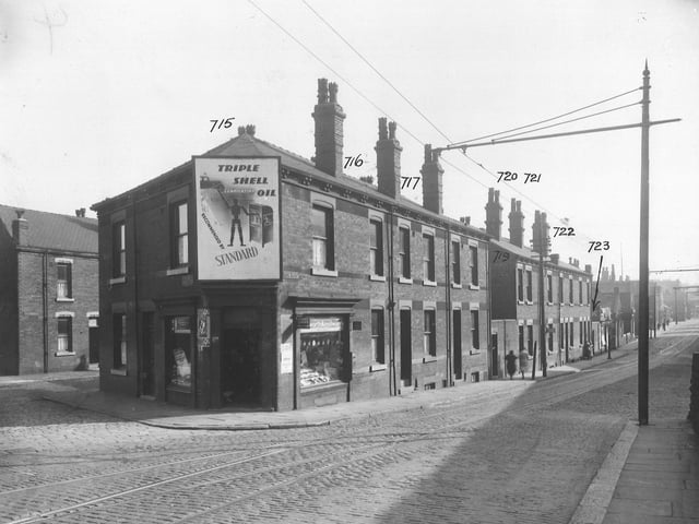 Balm Road's junction with Flax Mill Road; both cobbled roads with tram lines. A newsagent's is on the corner, with a large advertisement for Shell Oil over the door. Brick back-to-back houses with yards and communal toilet blocks. Small business premises and shops. On the right is a tall standard with an arm extending across the road, supporting power lines for trams. Pictured in September 1935.