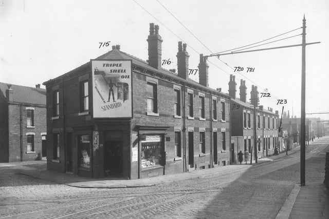 Balm Road's junction with Flax Mill Road; both cobbled roads with tram lines. A newsagent's is on the corner, with a large advertisement for Shell Oil over the door. Brick back-to-back houses with yards and communal toilet blocks. Small business premises and shops. On the right is a tall standard with an arm extending across the road, supporting power lines for trams. Pictured in September 1935.