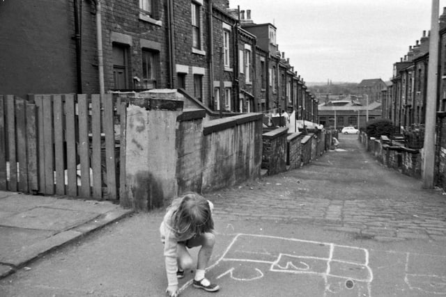 A young girl chalks out a grid for a game of hopscotch. The cobbled terraced Holderness Street behind her slopes down to Queen's Road at the bottom where the British Relay Depot can be seen. The large building dominating the background is St. Margaret's C of E Church on Cardigan Road.