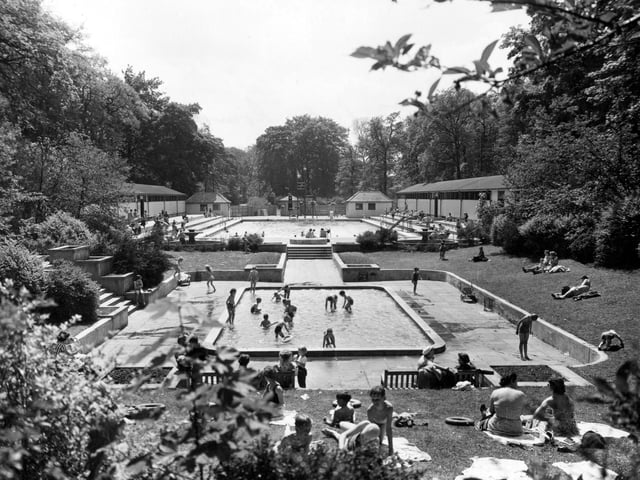Children playing in an open air swimming pool in Roundhay Park in June 1955. A shallow pool is in the foreground with a deeper pool behind. Parents sit on benches round the outside or lie on the grass. Changing rooms can be seen.