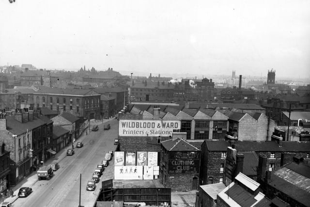 An  elevated view looking north along Meadow Lane towards Leeds Bridge and Hunslet Lane. In the foreground are the premises of Wildblood & Ward, printers, and number 57a, Leeds City Clothing Club. On the left is the Old George public house. Cars and lorries are parked on the road. The view extends to Leeds Parish Church and Quarry Hill Flats (both on the right in the distance). Advertising hoardings for chocolate, cigarettes and fruit juice are visible. Pictured in April 1955.