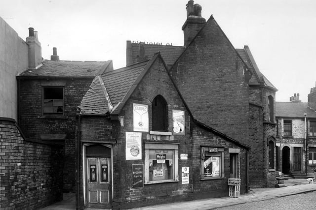 A disused shop on Holdsworth Street pictured in August 1958. A sign in the window advertises a Yorkshire Evening News article from November 1956, two years out of date. On the right edge, the entrance to Brookfield Place is visible.