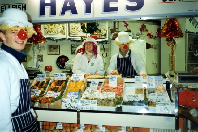 This photo thought to date from 1998, shows the staff of Thomas Hayes, a fishmongers at number 169 Kirkgate Market. The staff, in navy and white, have entered into the spirit of Christmas and are wearing colourful wigs and false noses. The counter display includes many varieties of fresh fish and shellfish. There are prawns, cockles, winkles, mussels, shrimp, crab claws and oysters as well as smoked fish such as sprats and finnan haddock.