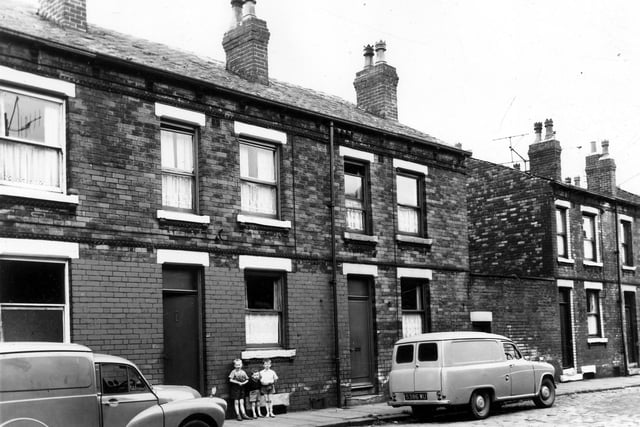 Back-to-back terraced houses separated between numbers 18 and 20 by a yard originally built to house the shared outside toilet. Two vans are parked in the street, are outside number 22 on the left and another, reg: 5386 WU outside number 20. Three boys in short trousers are stood under the window of number 22. This area was soon to be demolished and families relocated under a Leeds City Council slum clearance programme. Pictured in August 1963.