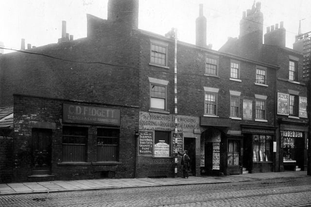 Neville Street and Francis Court in September 1910. On the left, number 12 premises of C.D. Fidgett cigar and tobacco manufacturer. Next right, number 13 with painted wall signs the Yorkshire Limewashing and Colouring Company. Workmen are measuring height of the building. The entrance to Francis Court can be seen, with entertainment posters on the walls. Number 14 is the property of Mrs. E.M. Hughes, dining rooms. There are two windows displaying services 'Dinners, Teas' and shop name. Number 15, extreme right, is L. Fisher, tobacconist.