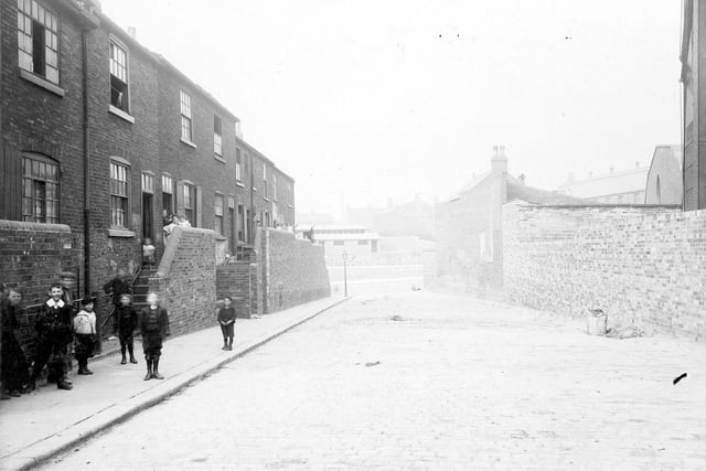 Row of brick dwellings in Quarry Hill, deemed to be in an unhealthy area. Natural wells in the area sometimes contaminated by raw sewage, leading to outbreaks of Cholera and other diseases. Water pipes have been installed in these houses and can be seen running to the side of the steps. The yard had High Street to one side and was bounded on the other by Leeds Corporation Gasworks, which is on right. Women and children are posing for the camera. Pictured in August 1906.