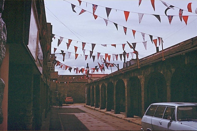 Bunting is strung across the Shambles in Wetherby with a patriotic red, white and blue theme and Union Jacks in celebration of the Silver Jubilee of Her Majesty Queen Elizabeth II. The old Market Shambles dates from circa 1811 and is designed with a colonnade, a row of columns supporting arches. It had been 10 butcher's shops. The Shambles converted to a Market Hall in 1888 and was paid for by public subscription.