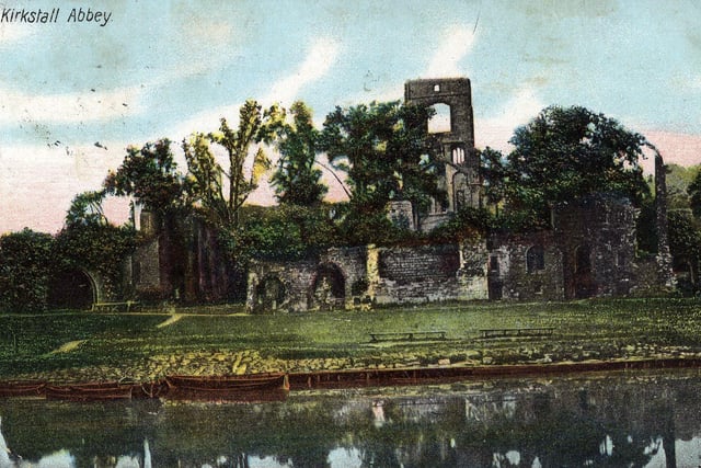 A tinted postcard view of Kirkstall Abbey from the River Aire. A few row boats are moored on the banks of the river. The postcard was mailed in Leeds in June 1906.
