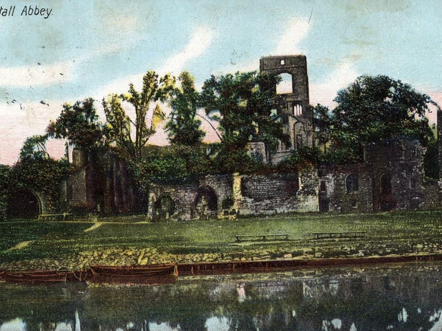 A tinted postcard view of Kirkstall Abbey from the River Aire. A few row boats are moored on the banks of the river. The postcard was mailed in Leeds in June 1906.