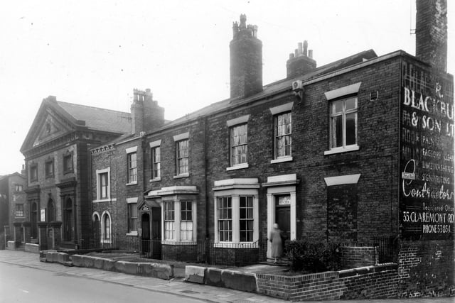 Burley Road in October 1959. To the left, at the corner with Olive Grove is the former Burley Lawn Methodist Chapel. It was being used as a print works by Walter Gardham Ltd, a notice on the door reads 'Rathmell and Oxley, Photolitho Ltd'. Moving right, number 211 Burley Road is a small house with one window to each floor. Next 213 has a porch with the name 'Dr Kelly Surgeon'. A woman is on the shop of 215, which has a notice over the door offering boot and shoe repairs.