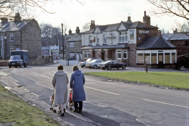 Looking south-east along Finkle Lane from Gildersome Green, showing The Junction public house on the right. Two women are pushing children in pushchairs in the foreground. The gable end of a terrace of houses on The Nooks is seen on the left.  Pictured in March 1984.
