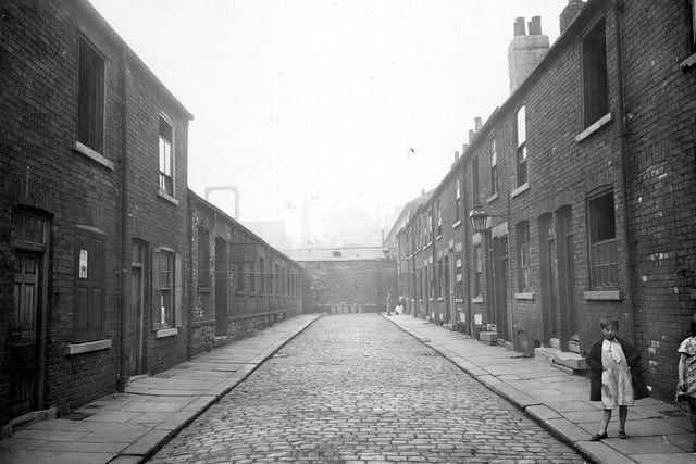 Rows of terraced houses on Cloth Street with some derelict with windows missing or boarded up. Children pose in the street. Dead end cobbled street with rows of dustbins at the end of the road. Pictured in August 1935.
