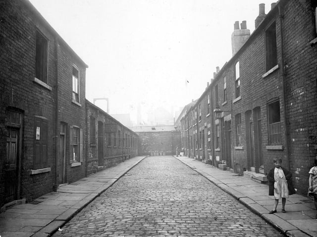 Rows of terraced houses on Cloth Street with some derelict with windows missing or boarded up. Children pose in the street. Dead end cobbled street with rows of dustbins at the end of the road. Pictured in August 1935.