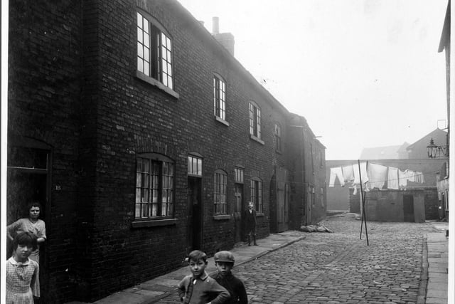 Showing Chadwick Court and various residential premises. In the background there is a line of washing hung across the road and in the foreground are several children and a woman in the doorway of number 15. Pictured in October 1935.