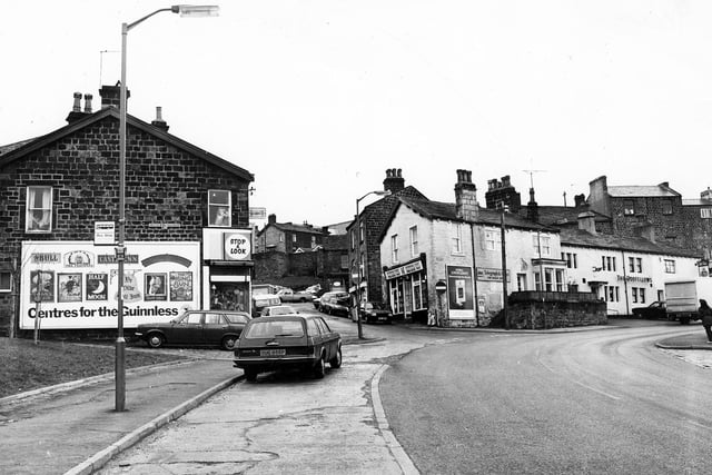 Yeadon's Kirk Lane, foreground, at the junction with The Green, right, and Old Haworth Lane; left. Town Street or 'The Steep' climbs up between the buildings in the centre to the High Street. At number 34 Town Street is Home Electrics with a large advertising campaign for Guinness on the gable end. The white building in the centre is a newsagents at the bottom of the steep. In the background, above the parked cars, buildings in Ivegate are visible. The two storey white building, right, is the Odd Fellows Public house or t'rag as it was known locally. It has been renamed The Rag and Louse in recent years.
