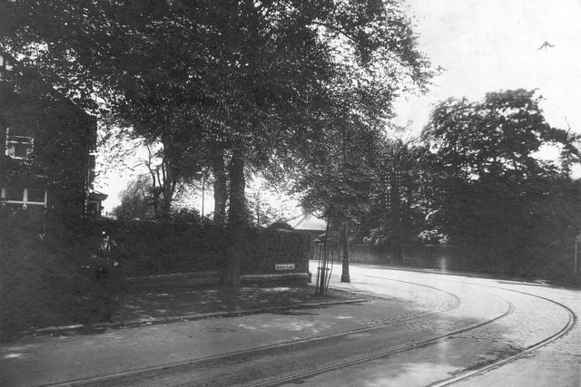 The junction of Harrogate Road and Street Lane shows on the left, a house, possibly the end of a terrace behind a high hedge. There are some people on the pavement including a man in the uniform of a transport company. A large mature tree overshadows the corner. Two sets of tram lines are seen on the road. In the right background is a stone wall behind which are more trees. Pictured in July 1931.