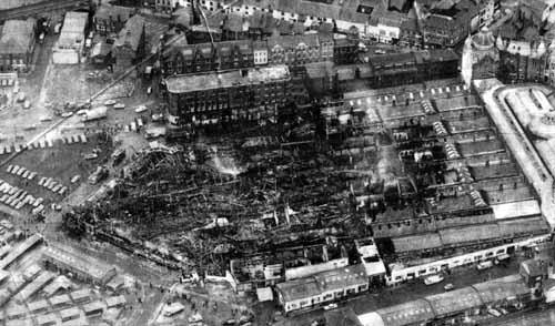 An aerial view of Kirkgate Market, looking south, in the aftermath of the fierce fire of Saturday, December 13, 1975. The photograph was taken the day after the fire and records the scene of devastation, as much of the market hall dating from 1857 was destroyed. The fire started and took hold only a half hour after closure to the public on a Saturday less than two weeks before Christmas. Draughts created by cross-ventilation between the stalls allowed the fire to escalate. The roof collapsed and a column of smoke hundreds of feet high hung over the city. Fortunately, the 100 firefighters attending the blaze managed to save the Vicar Lane end and the 1904 building. Many traders lost their Christmas stock and the day's takings.