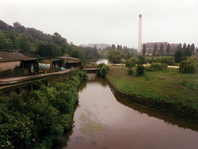 A view from the direction of Armley Mills towards Kirkstall Power Station, seen right. The Power Station was built between 1928 and 1930 by the City of Leeds and further extended between 1943 and 1944. It closed in October 1978 and the cooling towers were demolished in 1979. The remaining buildings were cleared by 1986. In the centre the weir is visible on the River Aire.  The trees on the left are part of Cunliff Wood.