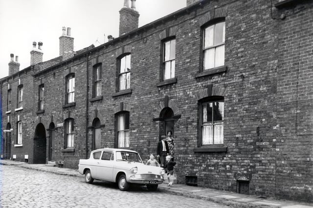 Back-to-back properties on Main Street, numbers run from 20 to 14, left to right with a passageway giving access to Parsonage View seen between numbers 20 and 18. Several people stand in the doorway of number 14 where a car is parked outside. Included in slum clearance plans for the Tong Road area of Armley. Pictured in August 1966.