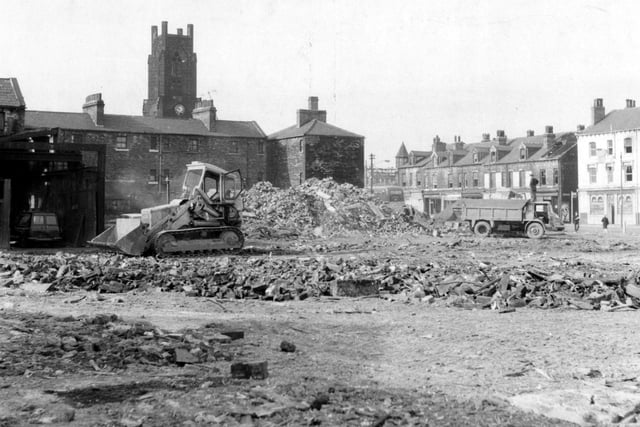 Demolition from Meadow Lane in Hunslet in February 1969. The view shows a digger in centre of a cleared area. A lorry is parked at the side and a church is visible in the background.