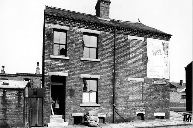 August 1950 and this photo shows number 3 Temple View Grove next to a derelict number 22 Chantrell Street. The front door to number 3 is open and a boy and a girl look at the camera from inside. Sandbags are piled up outside the house. Advertisements for Wisk washing powder and "our Gracie sings again" are visible on the side of number 22.