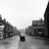 The view looking north along Brudenell Grove in November 1954. To the left of the photograph is a row of terraced shops, beginning at number 45 with 'J. Ashworth, boot repairs'. At number 47 is Edna Marfitt, drapers' and at number 49 is Fred Carr, television, radio and electrical installations'. There is a wide pavement which has puddles of rain and a wet looking surface. People walk along the pavement on each side of the road and cars are parked on the roadside. On the opposite side of the road are several rows of terraced houses, Brudenell View being in the foreground.