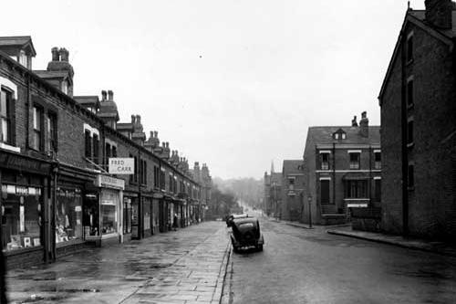 The view looking north along Brudenell Grove in November 1954. To the left of the photograph is a row of terraced shops, beginning at number 45 with 'J. Ashworth, boot repairs'. At number 47 is Edna Marfitt, drapers' and at number 49 is Fred Carr, television, radio and electrical installations'. There is a wide pavement which has puddles of rain and a wet looking surface. People walk along the pavement on each side of the road and cars are parked on the roadside. On the opposite side of the road are several rows of terraced houses, Brudenell View being in the foreground.