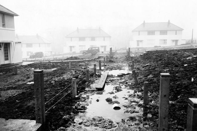 An unfinished, muddy footpath leading to Deanswood Drive on the Moortown Estate. A wire fence is on either side. The gardens are full of mud and cars are parked on the road. Pictured in February 1950.