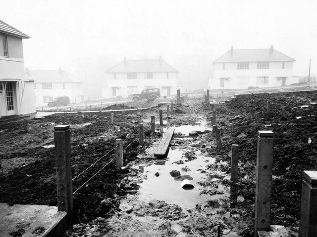 An unfinished, muddy footpath leading to Deanswood Drive on the Moortown Estate. A wire fence is on either side. The gardens are full of mud and cars are parked on the road. Pictured in February 1950.
