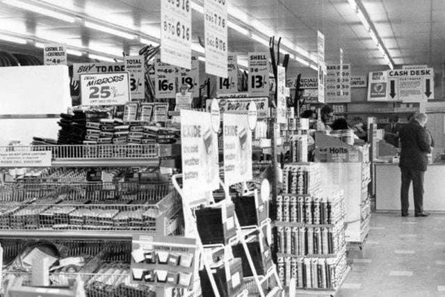Inside the new branch of Troy Tire & Auto Centre. It is arranged like a supermarket with motoring spares displayed on shelving in aisles in order for the customers to browse and select their own items. Prices are displayed overhead and these are all in pounds, shillings and pence as decimalisation was not introduced until 1971, the following year. At the far end of the shop a customer is paying for his goods at the cash desk.