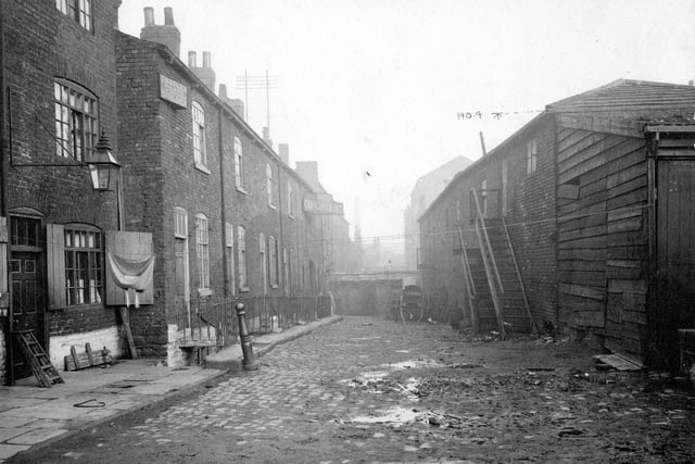 Black Swan Yard in Leeds city centre pictured in October 1898. Part of North Street Improvement Area, was number 2 North Street, now part of Vicar Lane. On the left houses, some with basements, access down steps from pavement. Railings act as barriers to open area. There are two wall signs for T. Butter, Coach builder. At the end of the yard are outside toilets and a cart. To the right, premises with wooden steps to upper doors. Yard has some cobbles, other parts unsurfaced.