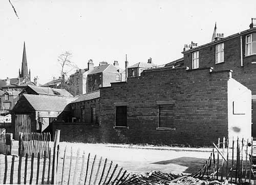 View looking across Fenton Place onto rear of garages on Back Ibbetson Place. Moving back, properties on Woodhouse Lane run from left to right. The spire on the left belongs to Trinity Congregational Church. The one just seen on the right is part of Blenheim Baptist Church both on Woodhouse Lane. Pictured in March 1966.
