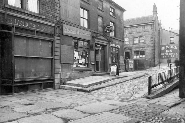Stocks Hill, Lower Town Street showing a block of stone built shop properties in September 1933. At the left edge is the butcher's shop belonging to Miss Isa Busfield (later to become Mrs Isa Croft) at number 145A, then Dunkirk Photographic Studios at number 147A, the business of James Blakey, 'Kiddies' portraits a speciality'. There is a large sign on the exterior wall that reads "Someone Somewhere Wants Your Photograph". This is followed by Leeds Permanent Building Society at number 147 with the pediment over the entrance.