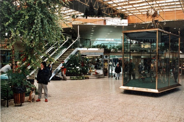 There is a glass case with an automaton with a clock on the top. Escalators lead up to the first floor and more shops. Many plants are visible in this view. There is a woman and a dog in the foreground.