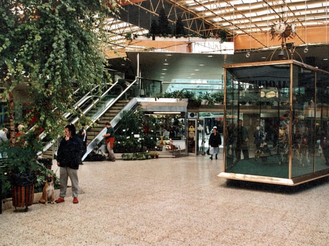 There is a glass case with an automaton with a clock on the top. Escalators lead up to the first floor and more shops. Many plants are visible in this view. There is a woman and a dog in the foreground.