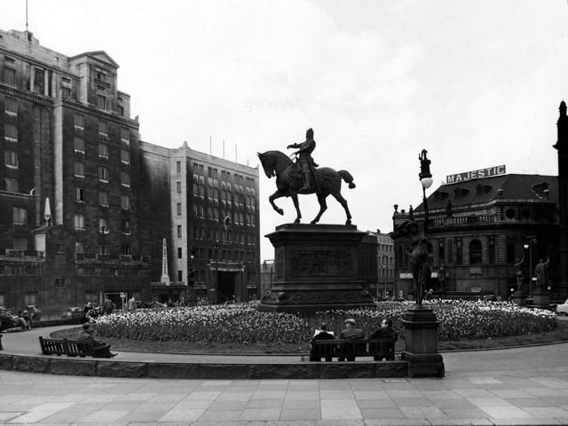People watch the world go by on benches in City Square in May 1955. The Majestic Theatre, the Queen's Hotel, the News Theatre and City Station are visible.
