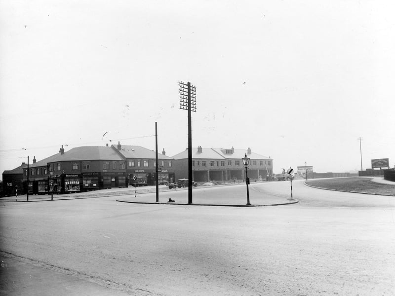 The junction of the Ring Road and Dewsbury Road. The island contains a gas streetlamp, two road signs, a multi-branched telegraph pole and an old black dog. In the background is a block of shops occupied and trading, with another block under construction. On the verge is a car and trailer. Men are unloading a lorry in the background.