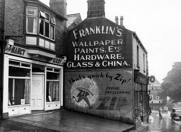 Shops on Stonegate Road pictured in August 1953. Nancy Kirby ladies hairdresser and Franklin's wallpaper, paints, etc. shown. Beckett Arms pub on the corner with Meanwood Road in the background. Advertisement for Zip dry cleaning on side of shop. People and cars visible.