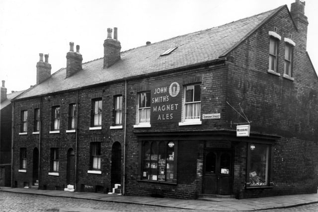 This view looks from Strawberry Avenue onto four properties on Dewhirst Street in May 1965. On the corner is an off-licence and confectioners with the name June Lily Bucktrout above the door. There is a painted sign for John Smiths Magnet Ales and a hanging advertisement for Woodbine cigarettes.