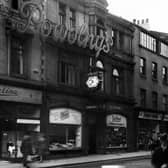 Bond Street, showing Powolny's Restaurant in the centre. The entrance to the restaurant has an awning, with a six sided clock above. The large neon sign "Powolny's" stretches across numbers 4 and 5 on the first floor. Number 4 is Martina, gown specialist, with displays of dresses in the windows. To the left of Powolney's entrance, a florist, to the right,Tetley, tobacco service. At number 6 on the right is Pearson & Denham, photographic material dealers. At the far left is a sign for Polly's Bar. The building has unusual decorative balconies at roof level below the gable ends, and shallow bay windows with curved glass above the entrance. In the foreground, a motorcycle and sidecar combination, and the rear of a Triumph convertible car. Pictured in June 1950.