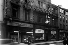 Bond Street, showing Powolny's Restaurant in the centre. The entrance to the restaurant has an awning, with a six sided clock above. The large neon sign "Powolny's" stretches across numbers 4 and 5 on the first floor. Number 4 is Martina, gown specialist, with displays of dresses in the windows. To the left of Powolney's entrance, a florist, to the right,Tetley, tobacco service. At number 6 on the right is Pearson & Denham, photographic material dealers. At the far left is a sign for Polly's Bar. The building has unusual decorative balconies at roof level below the gable ends, and shallow bay windows with curved glass above the entrance. In the foreground, a motorcycle and sidecar combination, and the rear of a Triumph convertible car. Pictured in June 1950.