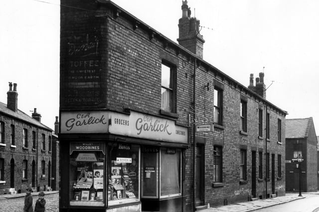 On the left edge of this view, properties on Main Street can be seen. Moving right onto Main Road, two boys stand next to a grocers and confectioners with the names C.& A. Garlick over the door. A painted wall sign above the window bears the name C. Briggs, the proprietor of a tobacconists here between the 1910s and the late 1940s. Pictured in  August 1966.