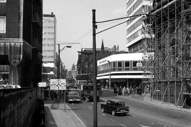 Park Row from Bishopgate Street in June 1967. A building covered in scaffolding is on the right. A restaurant and carpet store are behind. On the left is the Norwich Union Buildings and a tower block. Traffic signs are prominent in the foreground plus a sign to Leeds Bradford Airport.