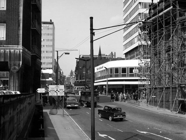 Park Row from Bishopgate Street in June 1967. A building covered in scaffolding is on the right. A restaurant and carpet store are behind. On the left is the Norwich Union Buildings and a tower block. Traffic signs are prominent in the foreground plus a sign to Leeds Bradford Airport.