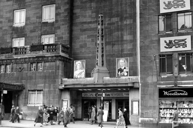 The front of the News Theatre on City Square in June 1953. Pictures bearing the portraits of Queen Elizabeth and the Duke of Edinburgh are visible either side of a column with 'The News Theatre' written down it. Above the doors is a sign saying 'Special Coronation Newsreel'. People are walking past. Next door is 'Woodlands' chemist.