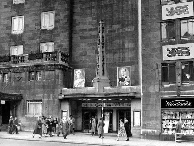 The front of the News Theatre on City Square in June 1953. Pictures bearing the portraits of Queen Elizabeth and the Duke of Edinburgh are visible either side of a column with 'The News Theatre' written down it. Above the doors is a sign saying 'Special Coronation Newsreel'. People are walking past. Next door is 'Woodlands' chemist.