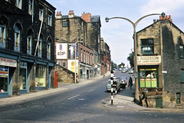 Chapel Hill from Morley Bottoms in August 1967. On the left hand side is the Conservative Club built in 1899 and three shops underneath. Beyond are the steps up to Bank Street and on to the Club fields of Cross Church Street Working Men's Club. After comes the cycle shop of John Hutchins and the base of the tall buildings which are also entered from Bank Street behind. Further up the hill are insurance offices and steps up to Bank Street. On the right hand side the photo shows the shop of Arthur Rogerson who took over the business of Baxter's fish, game and poultry merchants. This is the corner shop of a block that runs from 2 to 6 up Chapel Hill and 1 to 9 on Station Road. Some of the other buildings in this group had been demolished before they were listed.