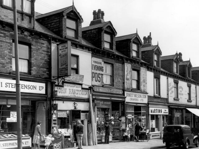A parade of shops in June 1965, from left, is I. Stephenson Ltd, butchers, then John Handyman Ltd, woodworkers supplies and 'The do-it-yourself Specialists'. Outside, two men look at a ladder, shelf brackets and colour charts for Dulux paints. Planks of wood lean next to the door. Next is J.W. Mullen's, newsagents, which also sells toys. A scooter hangs in the window and a magazine rack is visible in the entrance. Continuing right is Earnest Wilson & Son (Drapers) Ltd, then Martins the Cleaners, part of a chain of shops. On the right is C.W. Christopher & Son, plumbers & sanitary engineers.