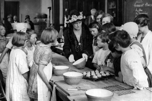 The Lady Mayoress, Ella Lupton, attends a lesson in cookery at Blenheim Council School on Blenheim Walk in July 1927. The lesson for this particular occasion is 'Coconut Buns' and the recipe has been chalked up on the blackboard, far right, for the children to follow. The Lady Mayoress appears to be discussing the ingredients and method with the pupils.
