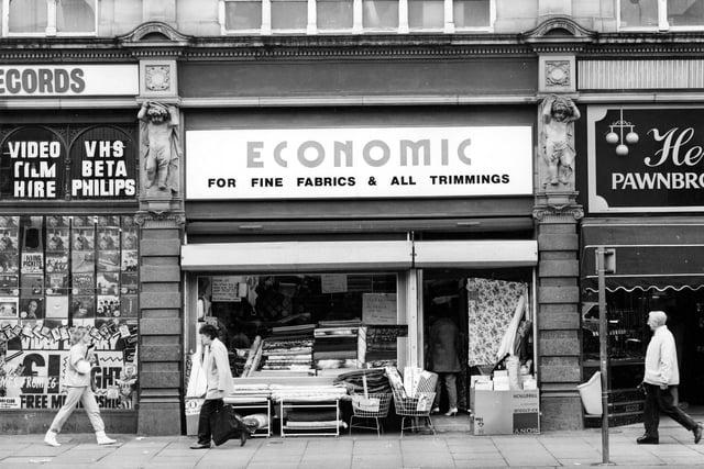 Market Buildings from Vicar Lane, with Economic Woollen Company, fabrics in the centre. Various fabrics can be seen on display in the window and outside the shop. On the left Cut Price Records has a display of records in the window. Joseph Hemsworth, jeweller is on the right. Pictured in May 1984.